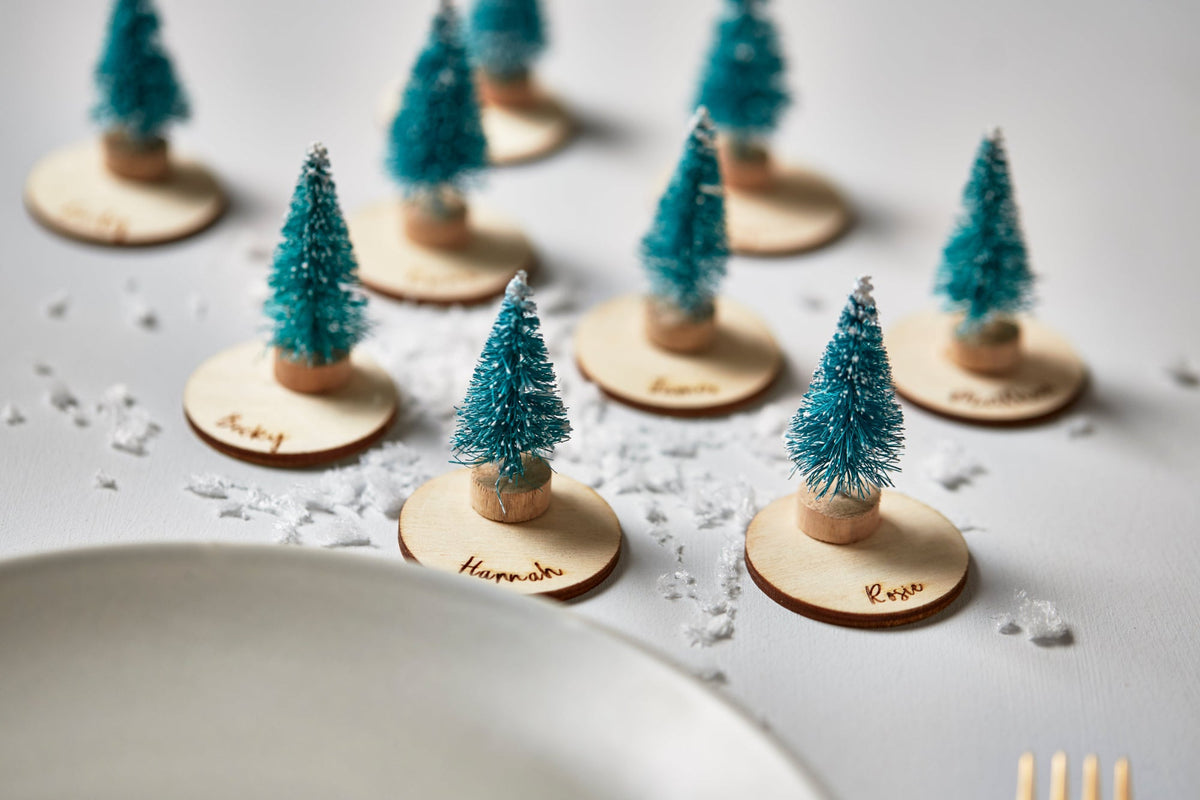 A set of miniature Christmas tree place cards on round wooden bases, styled on a white table with scattered faux snow and a white plate in the foreground.