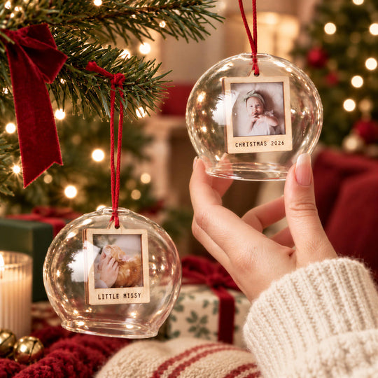 A hand holding two glass ornaments with photos of a baby, surrounded by a Christmas tree and other holiday decorations.