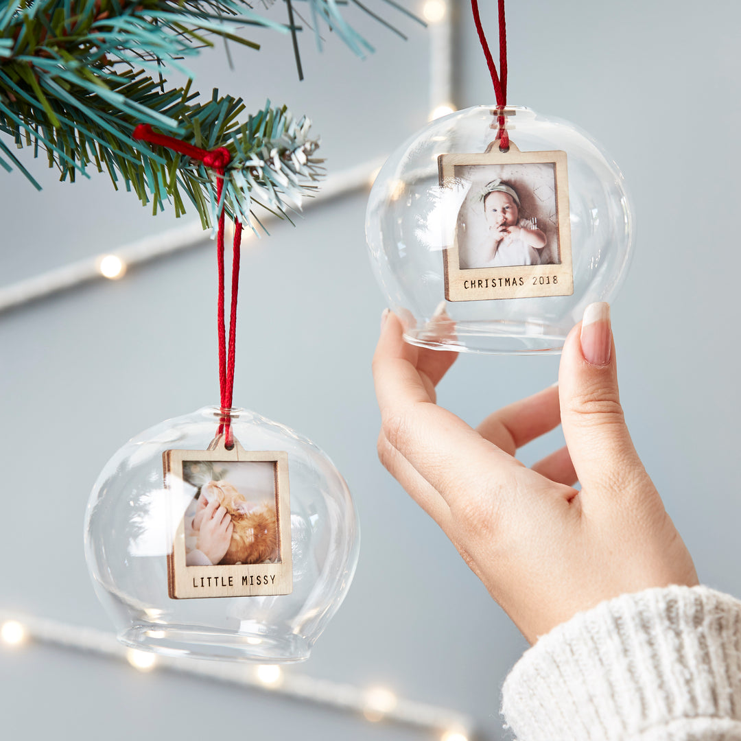 A hand holding two glass ornaments with photos of a baby, hanging from a Christmas tree.