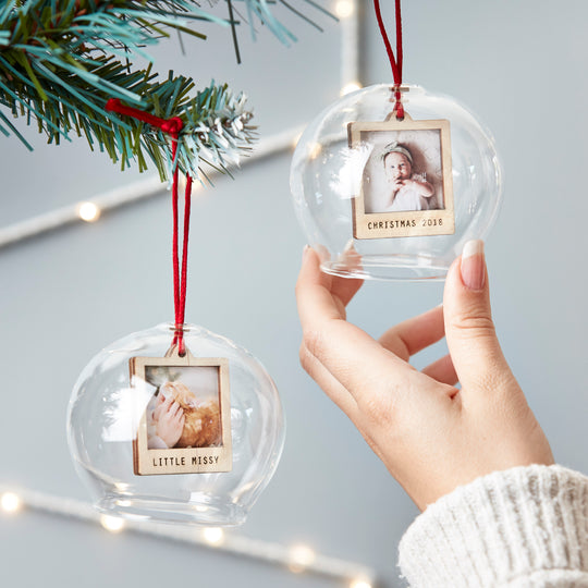 A hand holding two glass ornaments with photos of a baby, hanging from a Christmas tree.