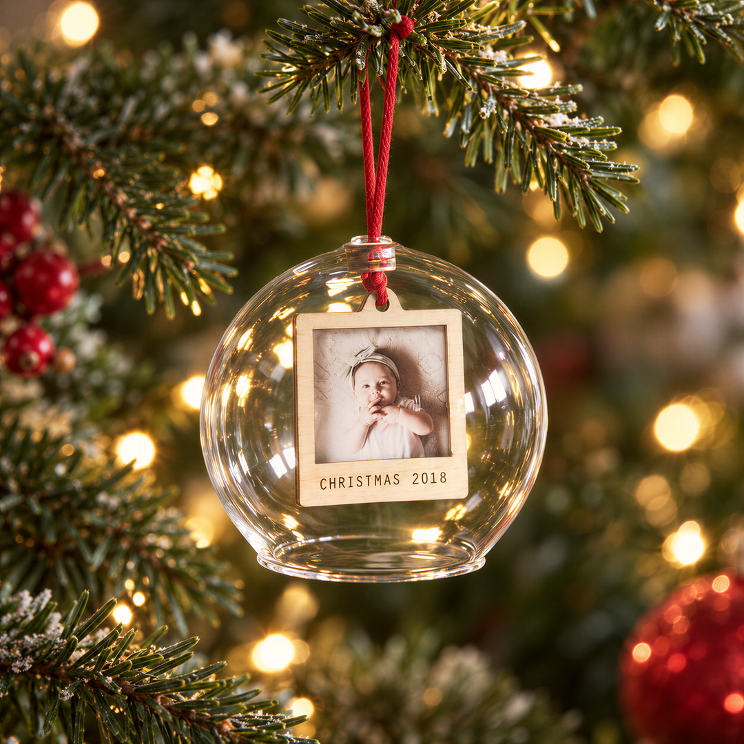 A glass ornament with a photo of a baby hangs from a Christmas tree, surrounded by twinkling lights and red ornaments.