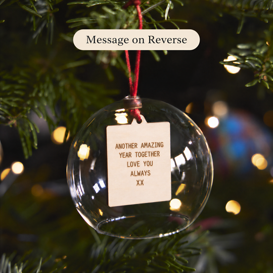 A clear glass ornament with a wooden tag attached, hanging on a Christmas tree.