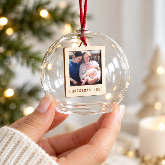 A hand holding a glass ornament with a photo of two people inside, surrounded by holiday decorations such as a Christmas tree and a candle.