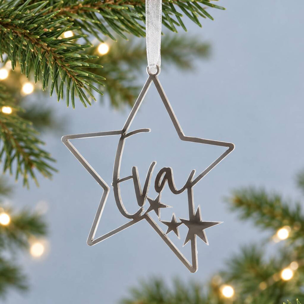 A silver star-shaped Christmas ornament with the name 'Eva' cut out in the center, hanging from a white ribbon on a pine tree branch with warm string lights in the background.