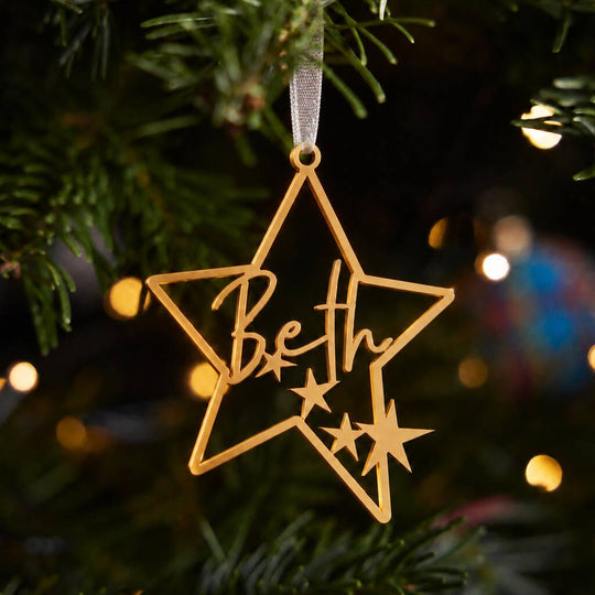 A gold star-shaped Christmas ornament hanging from a green Christmas tree branch, with the name &