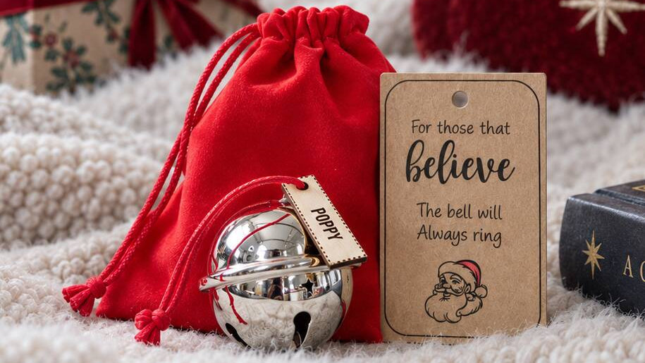 A festive Christmas gift set featuring a red drawstring pouch, a silver bell, and a personalized kraft paper tag that reads 'For those that believe The bell will Always ring'. The scene is styled on a soft white blanket with a wrapped gift box and a red stocking in the background.